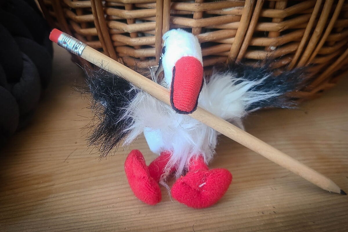vor einem Korb sitzt ein sehr kleiner Plüschstorch mit einem Bleistift unter dem Schnabel