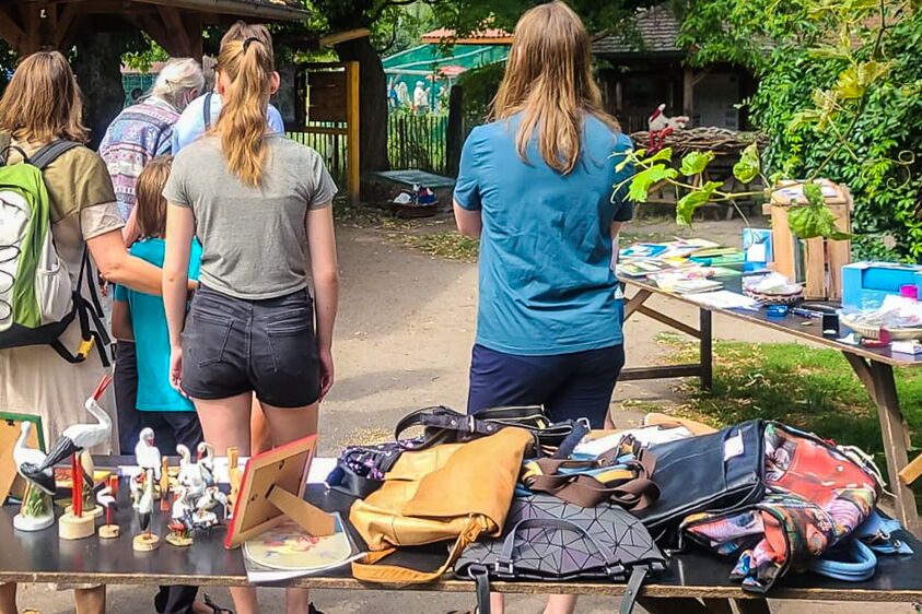einige Gäste sind von hinten zu sehen, davor Tische mit diversen Störchen und Taschen